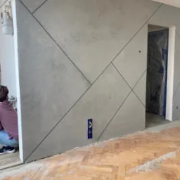 Ściana pokryta szarą gładzią z geometrycznym wzorem z ciemnych linii, widoczny fragment pracownika, zabezpieczone folią drzwi i parkiet przed cyklinowaniem.