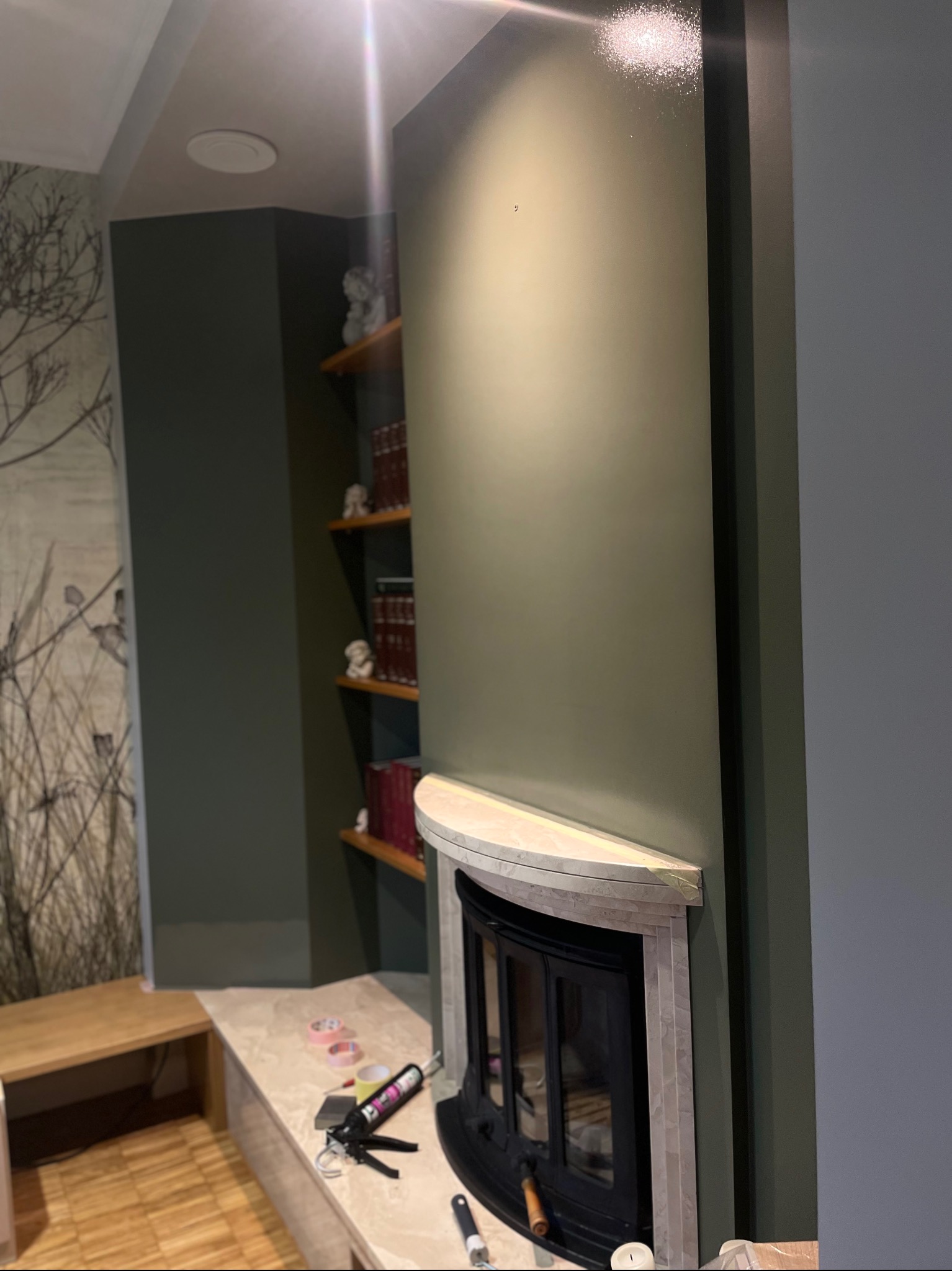 Montaż obudowy kominka z marmurowymi elementami, widoczne narzędzia i materiały budowlane, ściana pomalowana na oliwkowy kolor, półki z książkami w tle.