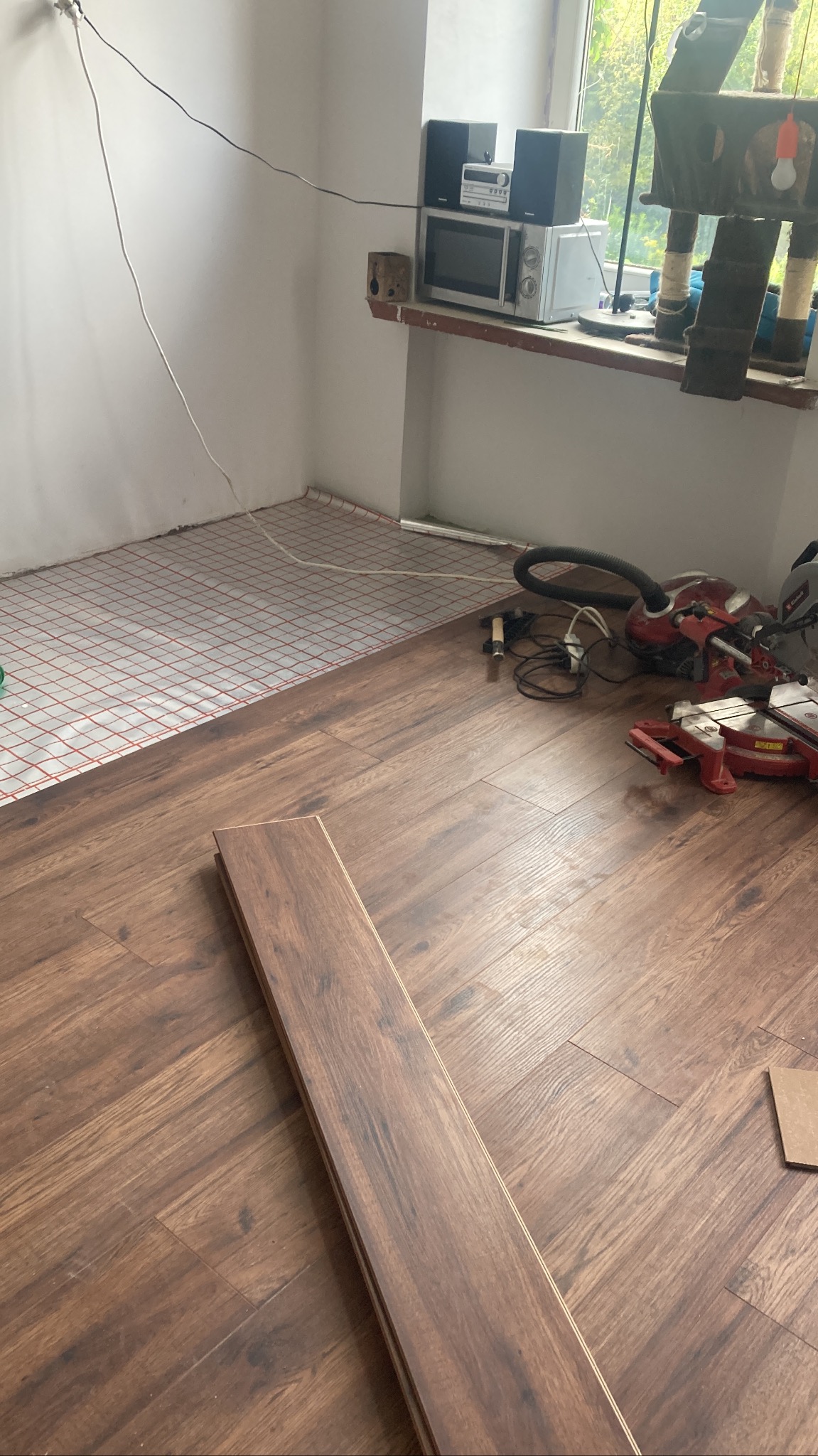 Proces układania paneli podłogowych w pokoju, widoczne panele o ciemnym odcieniu drewna, mata pod panele, narzędzia pracy w tle oraz sprzęt AGD na parapecie okiennym.