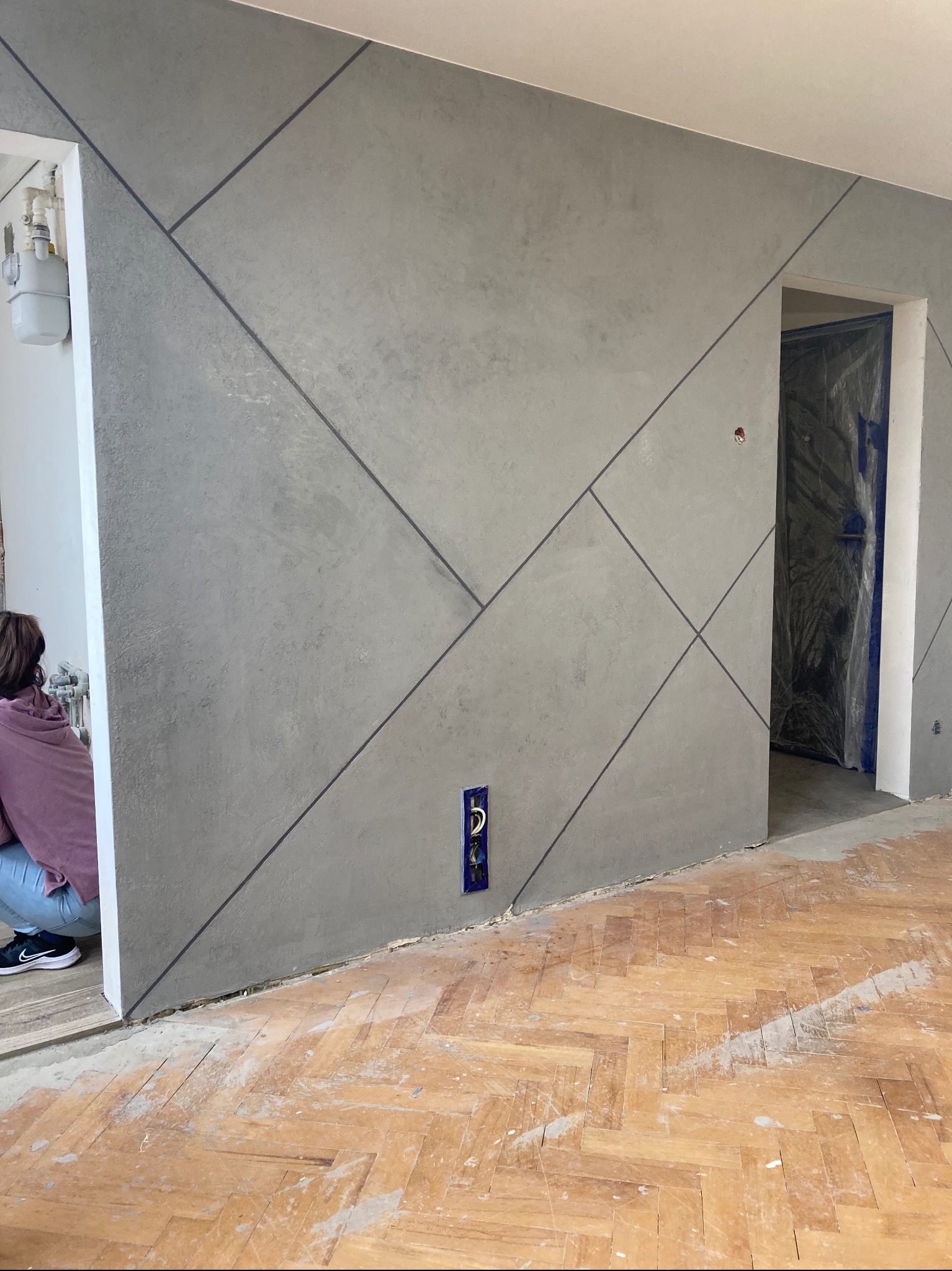 Ściana pokryta szarą gładzią z geometrycznym wzorem z ciemnych linii, widoczny fragment pracownika, zabezpieczone folią drzwi i parkiet przed cyklinowaniem.