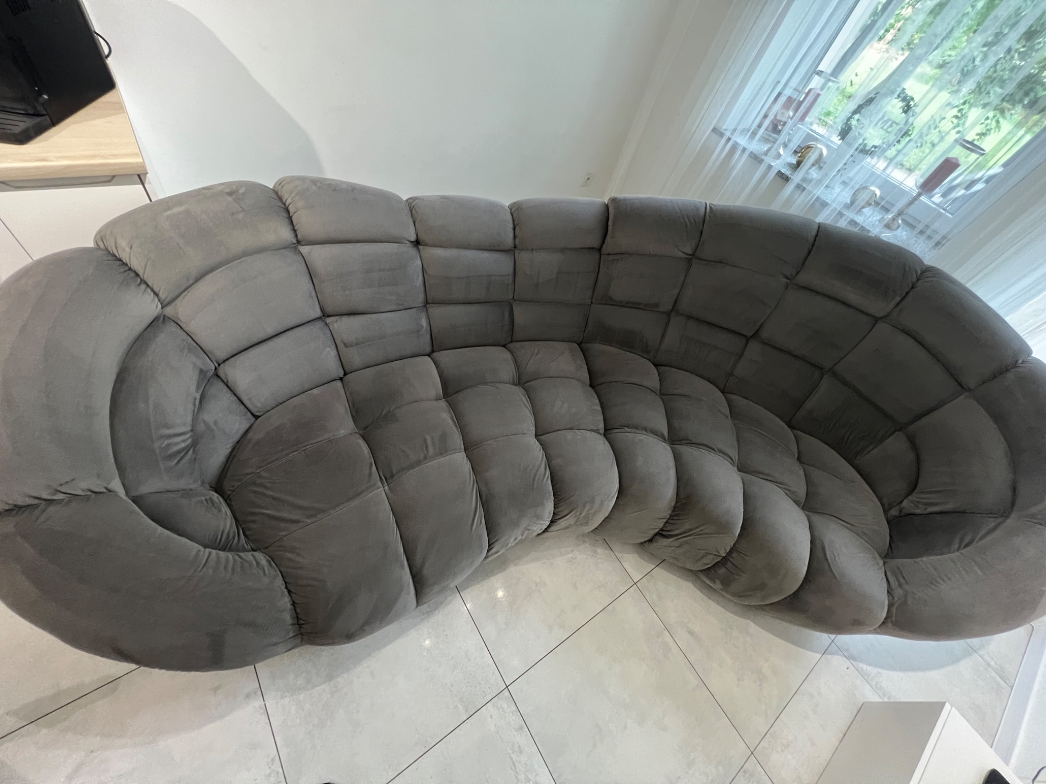 Narożna sofa tapicerowana w szarym odcieniu, z wyraźnymi podziałami na kwadratowe segmenty, ustawiona w jasnym wnętrzu z białymi kafelkami na podłodze i oknem z delikatnymi firankami w tle.