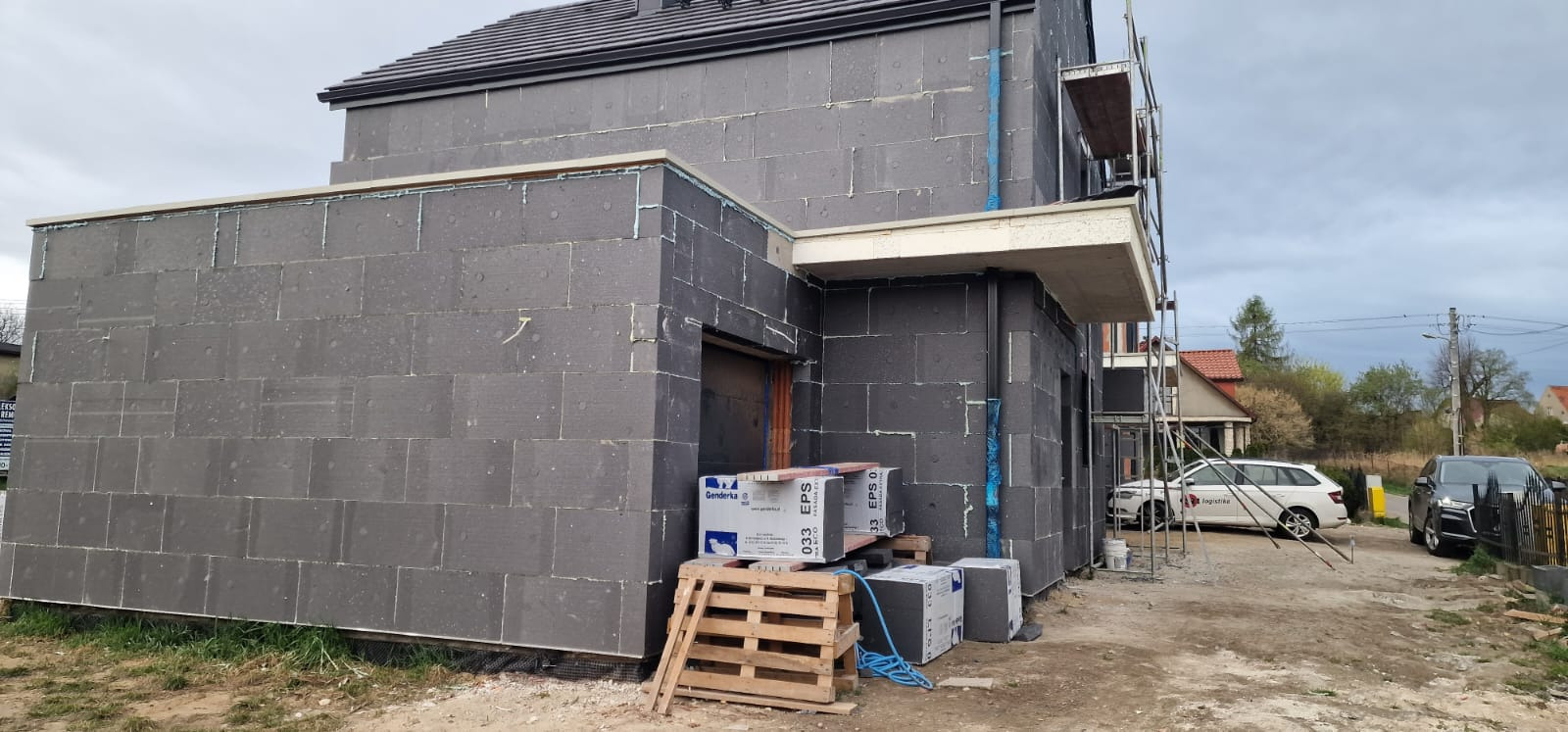 Dom w trakcie ocieplania: szare płyty styropianowe na elewacji, rusztowania, paleta z materiałami budowlanymi, okna w stanie surowym. Widok z zewnątrz.