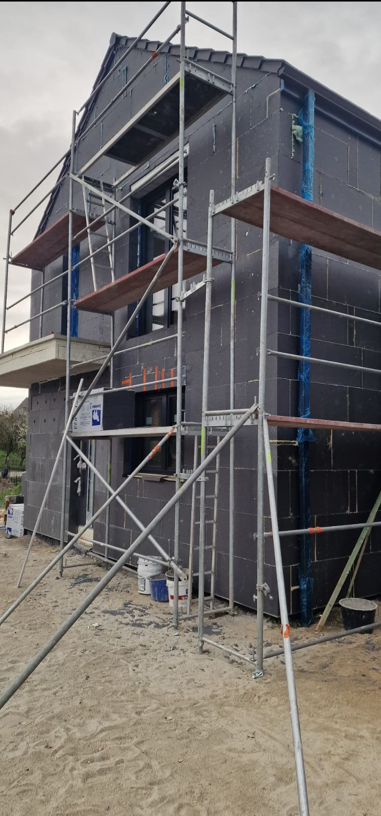 Dom w trakcie ocieplania, widoczne czarne płyty izolacyjne na elewacji i rusztowanie. Fragment budynku z oknami, w tle drzewa i pochmurne niebo.