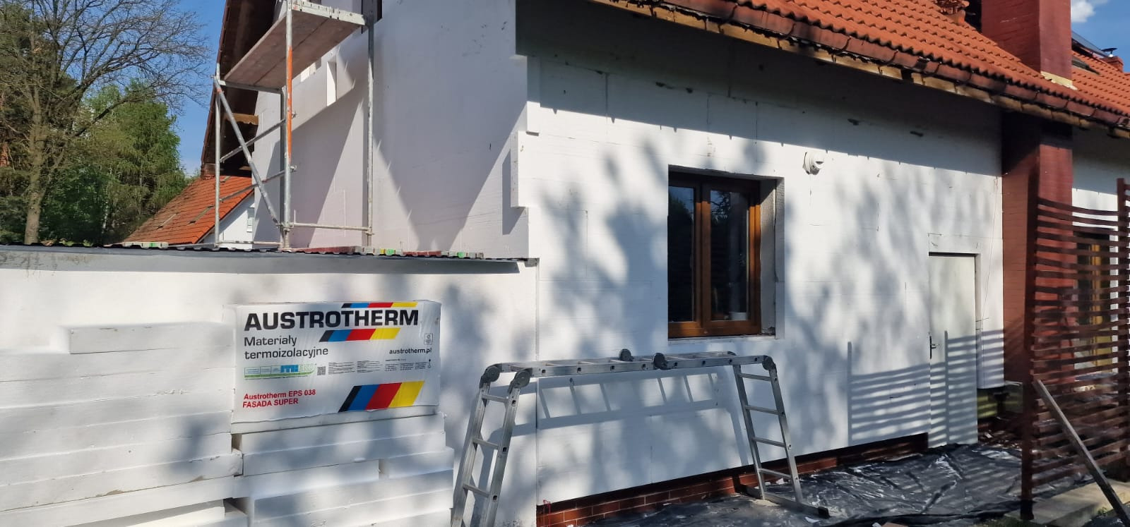 Dom w trakcie ocieplania, widoczne płyty styropianowe Austrotherm, rusztowanie i drabina oparta o ścianę. Częściowo ocieplona elewacja budynku jednorodzinnego.