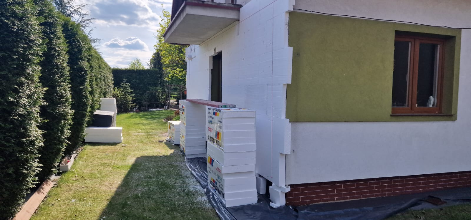Dom w trakcie ocieplania: biały styropian, zielona ściana z oknem, ceglana podmurówka. Częściowo ocieplona elewacja, materiały budowlane na trawie.