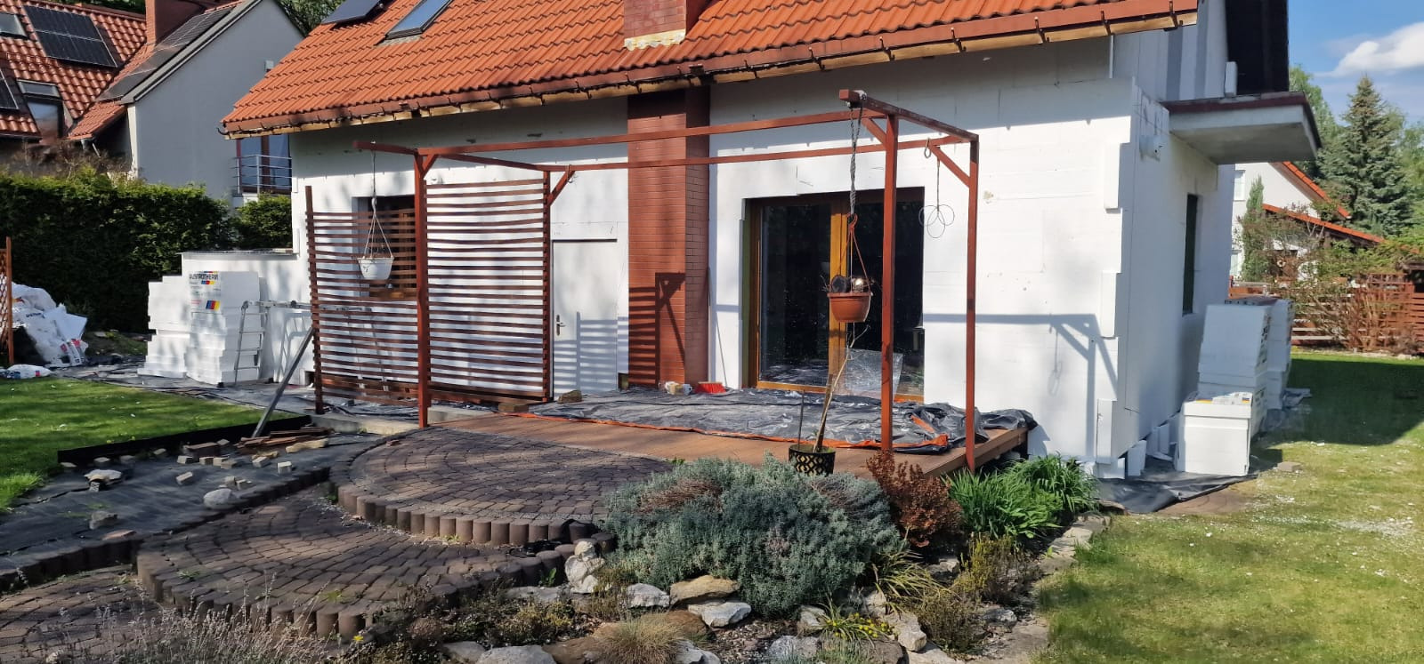 Dom w trakcie ocieplania, widoczne białe płyty izolacyjne, drewniana pergola i elementy dekoracyjne ogrodu. Prace elewacyjne w toku.