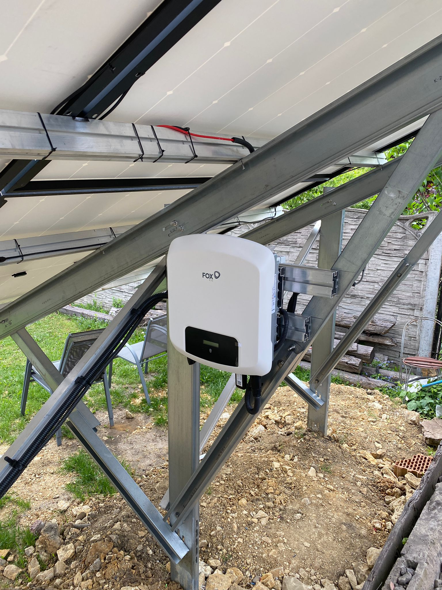 Biały falownik FOX pod panelami fotowoltaicznymi, zamontowany na metalowej konstrukcji. Widoczne kable, elementy montażowe i fragment ogrodu w tle.