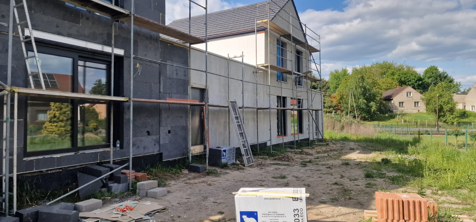 Dom w trakcie ocieplania, widoczne rusztowania, styropian grafitowy i biały. Część elewacji wykończona. Okna w czarnych ramach. Prace w toku.