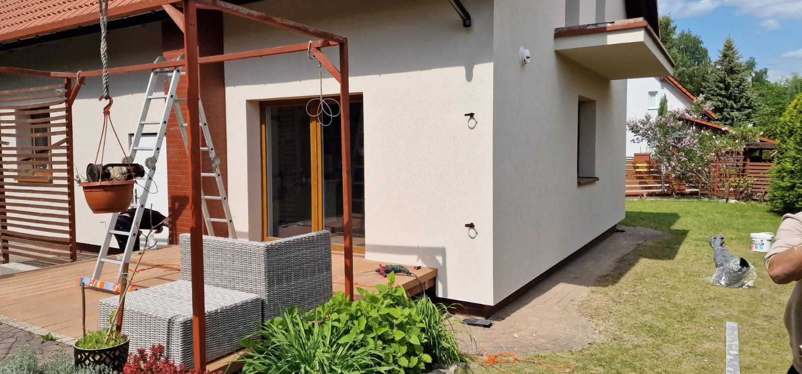 Jasna elewacja domu jednorodzinnego z elementami dekoracyjnymi i oknem. Widoczne narzędzia i drabina sugerują prace wykończeniowe.