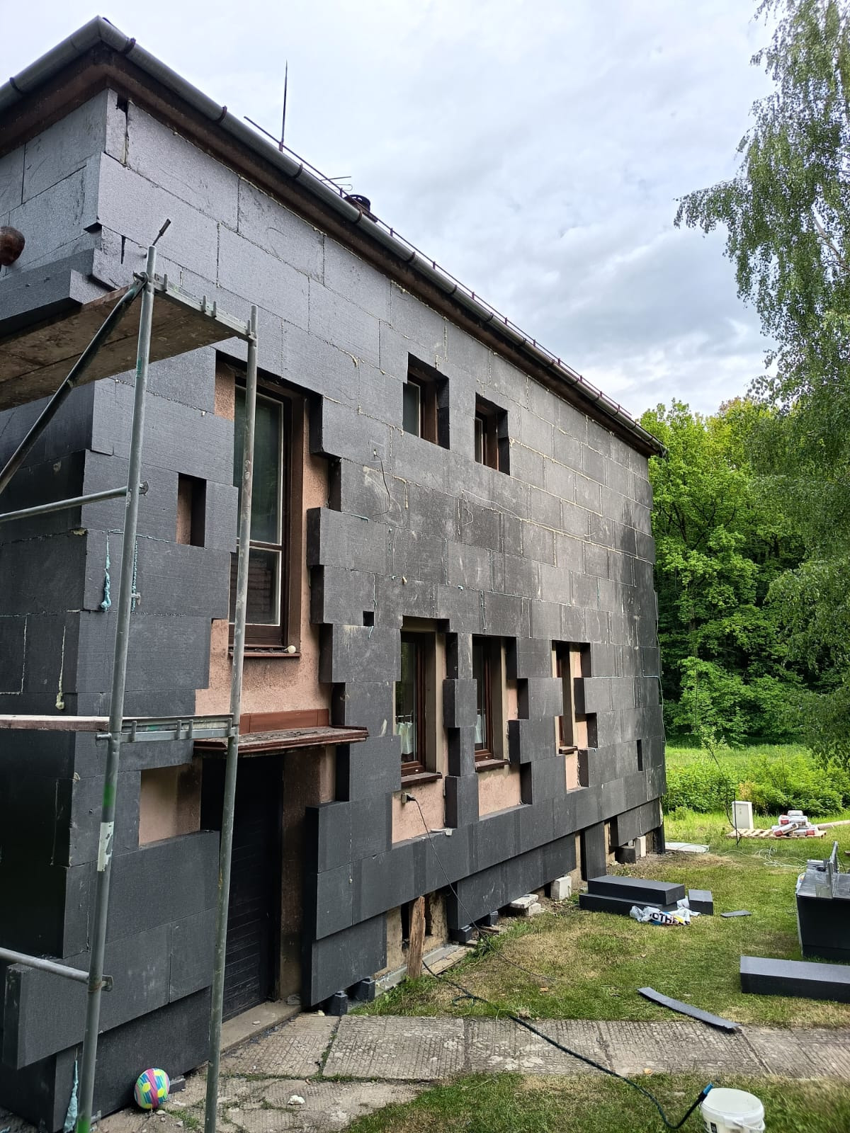 Dom w trakcie ocieplania szarą wełną mineralną. Widoczne rusztowanie, fragmenty elewacji bez izolacji, zielone otoczenie i fragment nieba.