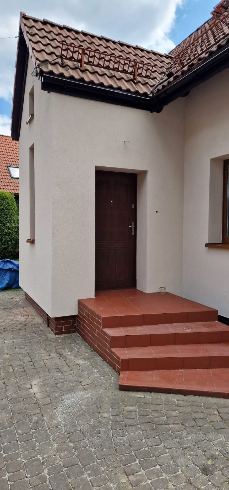 Odnowiona elewacja domu z brązowymi płytkami na schodach wejściowych i cokole. Jasna elewacja kontrastuje z ciemnym dachem i stolarką okienną.