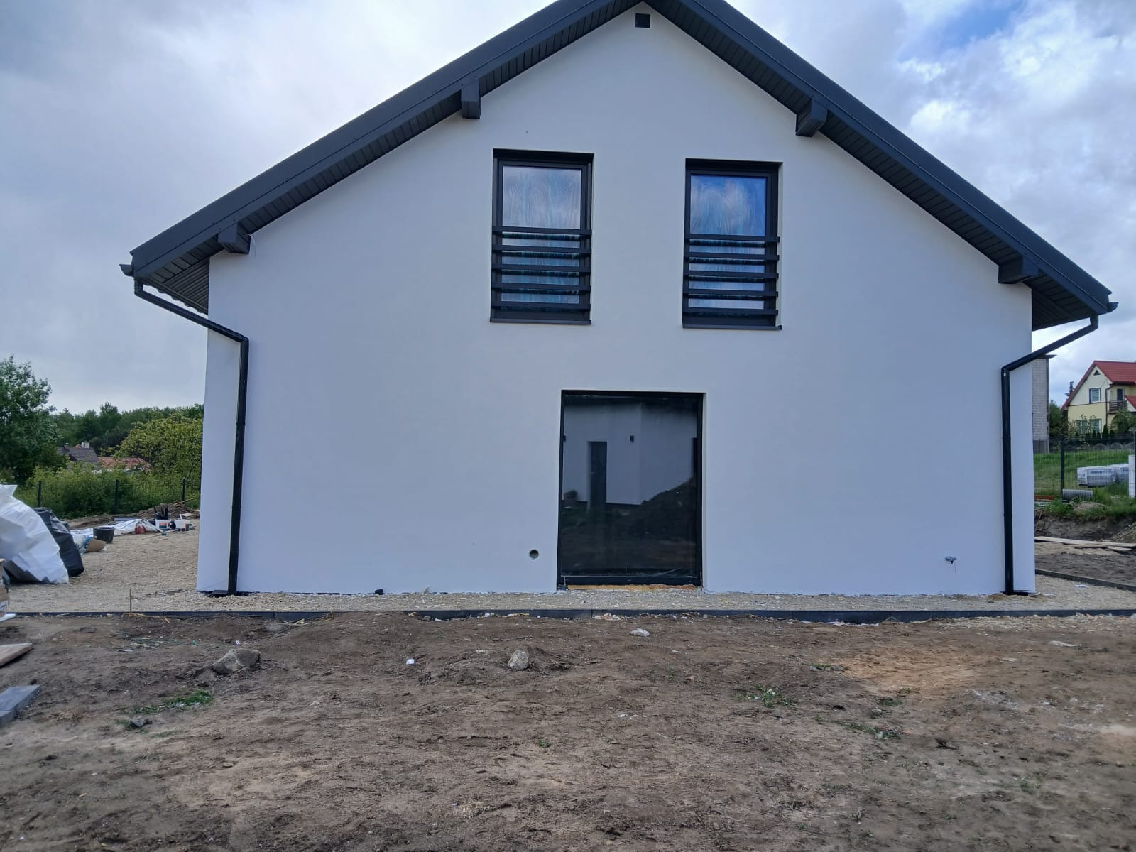 Biały dom jednorodzinny z ciemną stolarką okienną i dachem, świeżo po wykonaniu elewacji, widok z przodu na tle pochmurnego nieba. Minimalistyczny design.