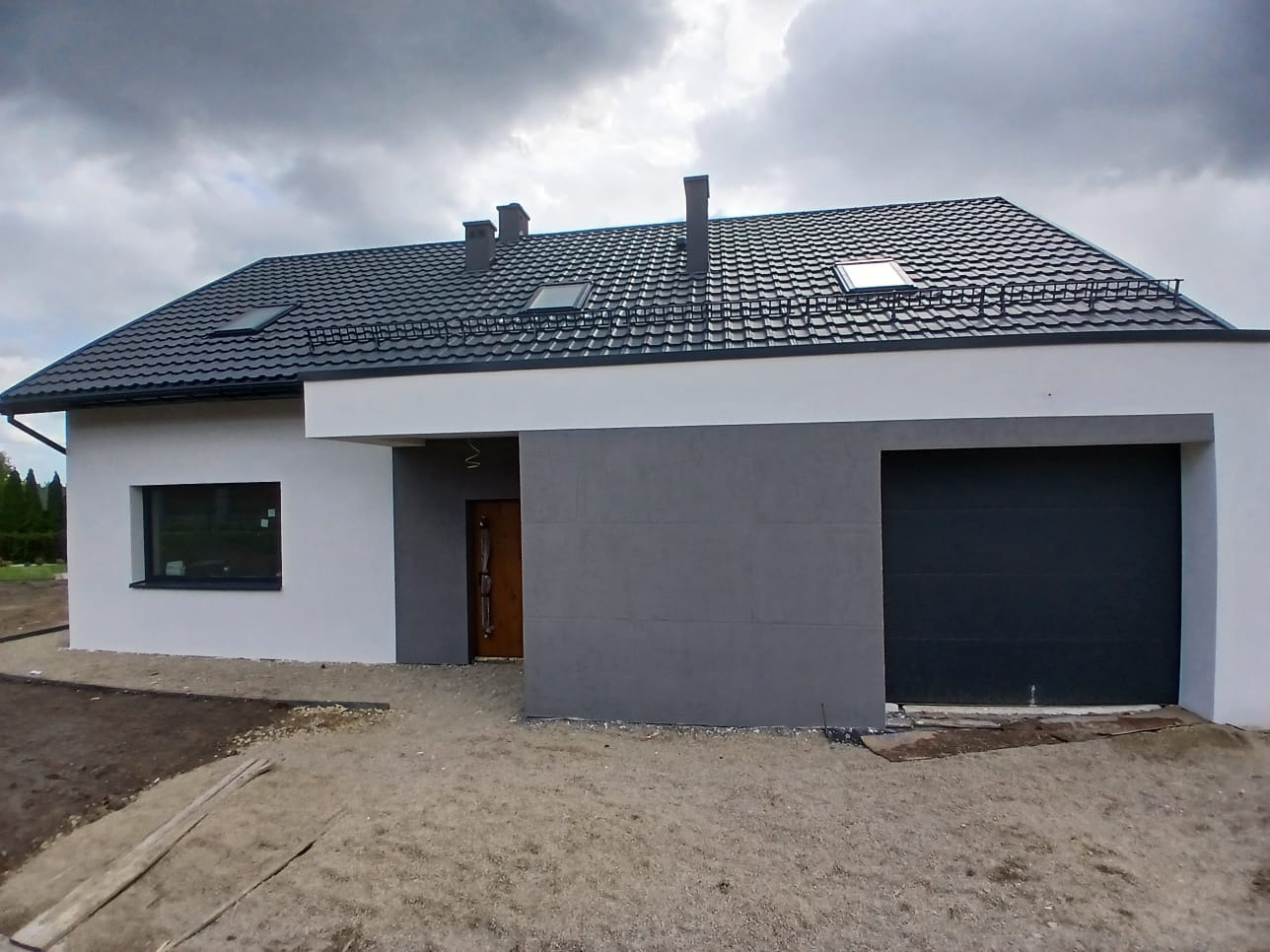 Nowoczesny dom jednorodzinny z szarą elewacją, ciemną dachówką i garażem. Widoczne okna dachowe i fragment ogrodu w pochmurny dzień.