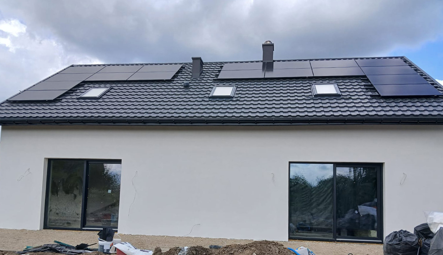 Nowoczesny dom z panelami fotowoltaicznymi na grafitowym dachu. Biała elewacja, okna z czarnymi ramami. Ujęcie z dołu, widoczne fragmenty gruntu.