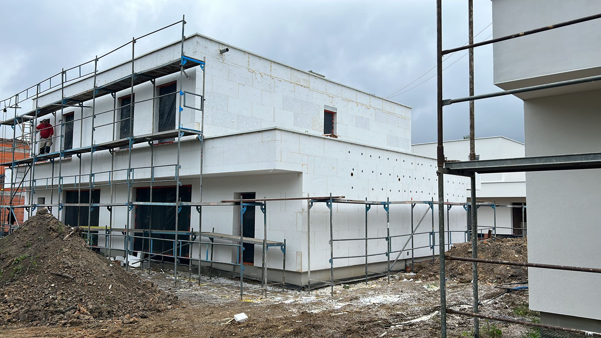 Ocieplenie budynku mieszkalnego styropianem, widoczne rusztowania i pracownik na tle pochmurnego nieba. Fragment ocieplonej elewacji sąsiedniego budynku.