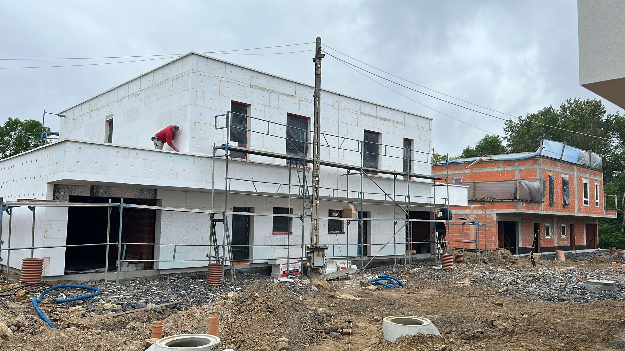 Dwa domy w trakcie budowy: jeden ocieplany styropianem, drugi z czerwonej cegły. Na pierwszym domu pracownik na rusztowaniu. Widoczne materiały budowlane.