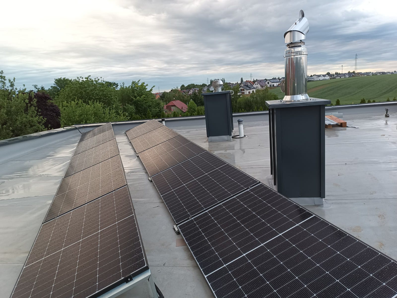 Instalacja paneli fotowoltaicznych na płaskim dachu z kominem i widokiem na okoliczne domy. Panele są czyste, po deszczu. Widoczne elementy montażowe.