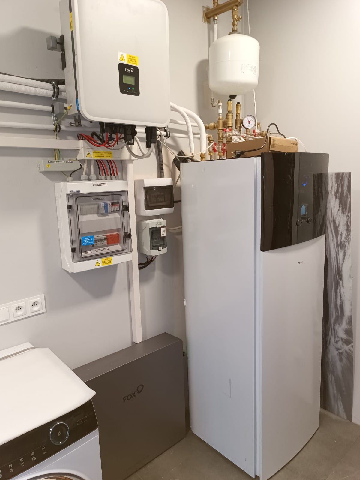 Biała pompa ciepła Fox-Ess z zbiornikiem wyrównawczym i rozdzielnicą elektryczną na ścianie w pomieszczeniu gospodarczym. Widoczna pralka.