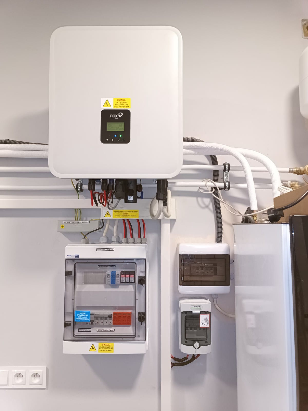 Biały inwerter FoxESS z rozdzielnicą PV AC i okablowaniem na jasnej ścianie. Widoczne elementy instalacji fotowoltaicznej, zabezpieczenia i przyłącza.