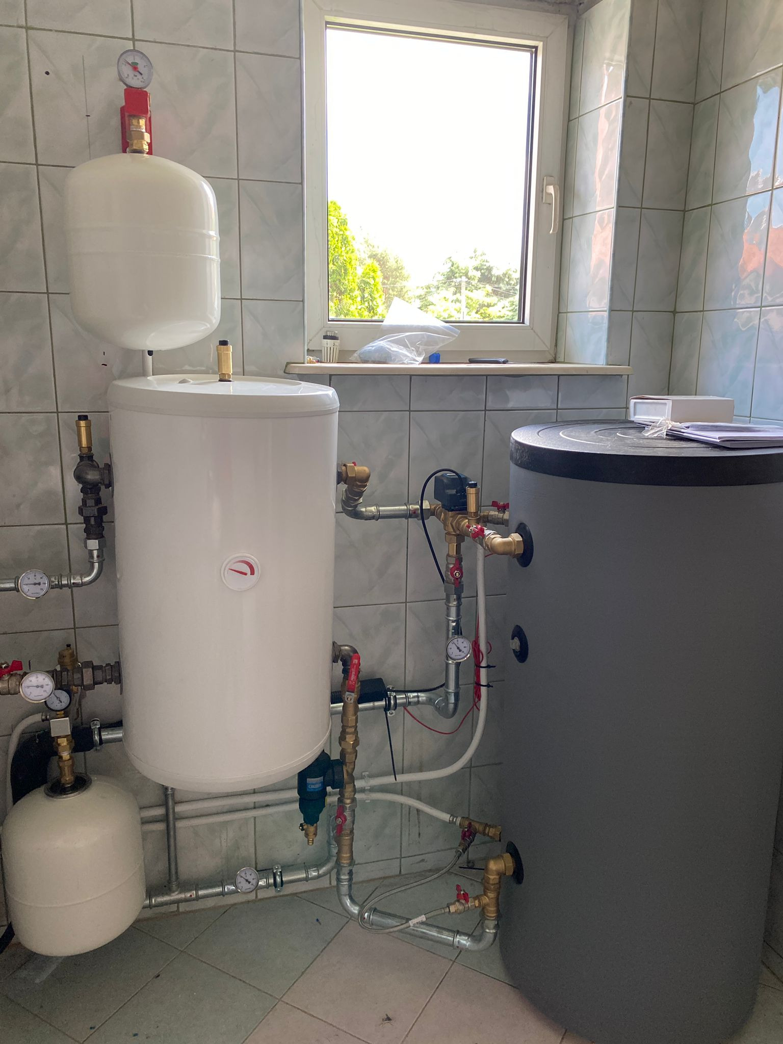 Biały zasobnik ciepłej wody użytkowej z osprzętem, w kotłowni z oknem i szarą płytką ścienną. Widoczne liczne zawory, manometry i rury.