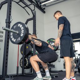 SlimBody Rzeszów - Mężczyzna wykonuje przysiad ze sztangą na stojaku pod okiem trenera w siłowni, widoczne tatuaże na ciele, podłoga wyłożona sztuczną trawą, na obciążeniach sztangi napis PROUD.