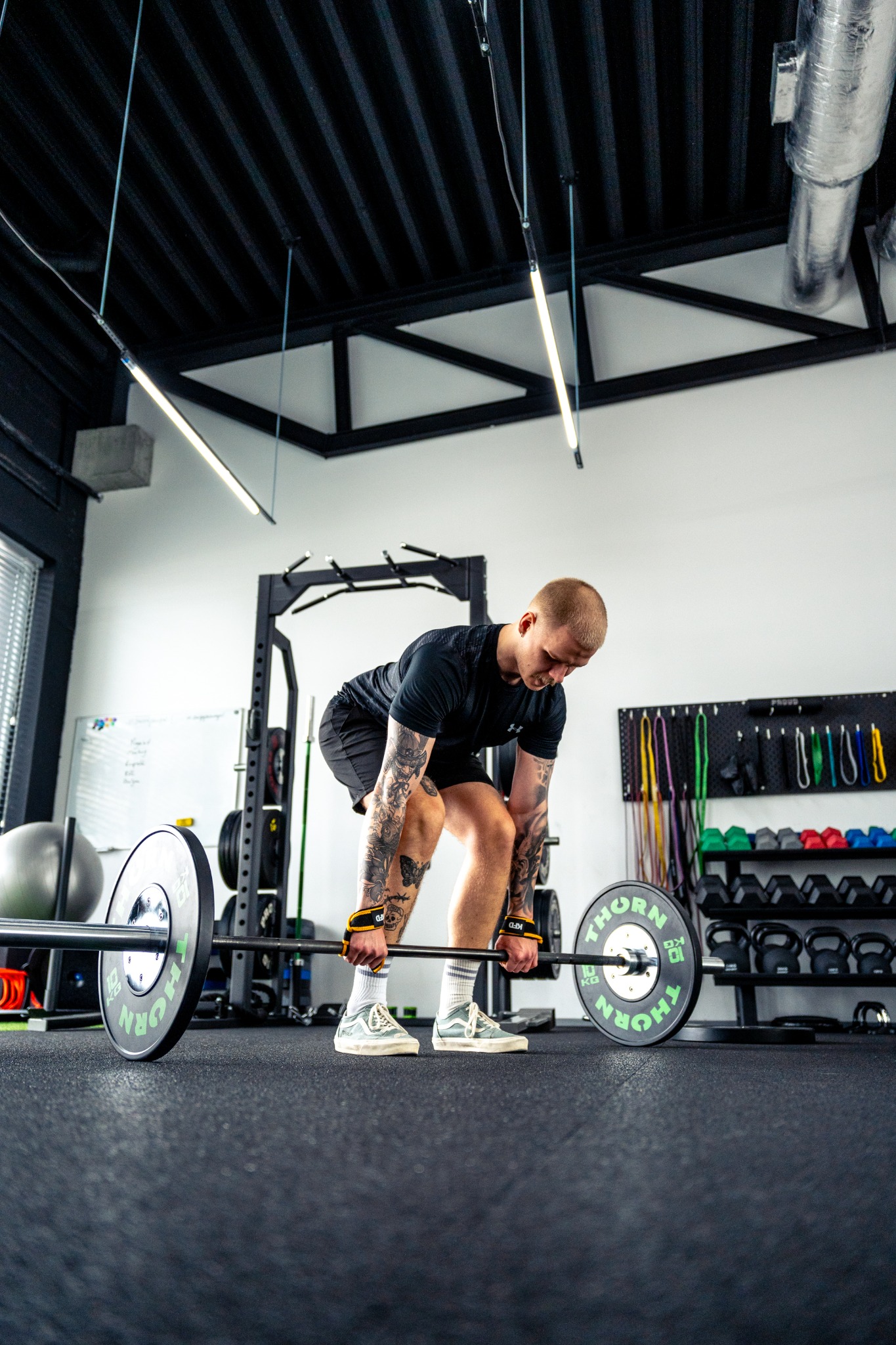 Mężczyzna z tatuażami na rękach przygotowuje się do podnoszenia sztangi w nowoczesnej siłowni, widoczne kettlebells i inne akcesoria do ćwiczeń.