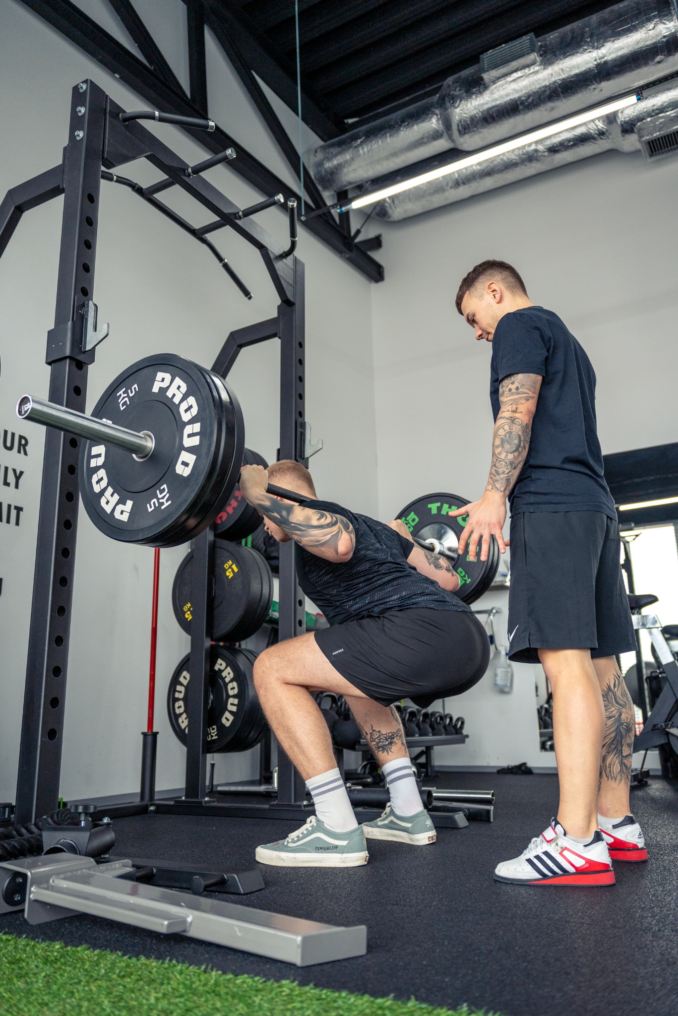Mężczyzna wykonuje przysiad ze sztangą na stojaku pod okiem trenera w siłowni, widoczne tatuaże na ciele, podłoga wyłożona sztuczną trawą, na obciążeniach sztangi napis PROUD.