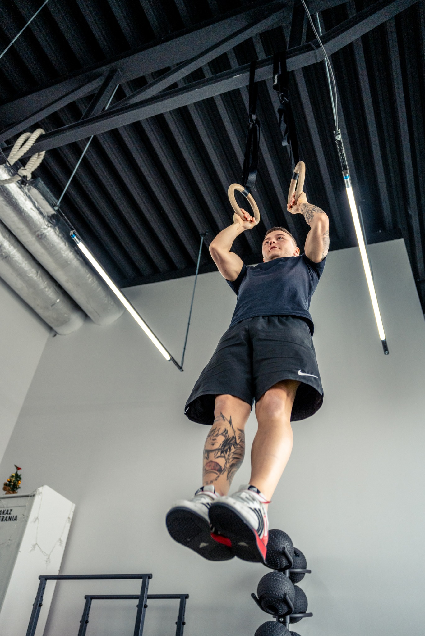 Mężczyzna z tatuażami na rękach i nogach ćwiczy na kółkach gimnastycznych w siłowni, widok z dołu.