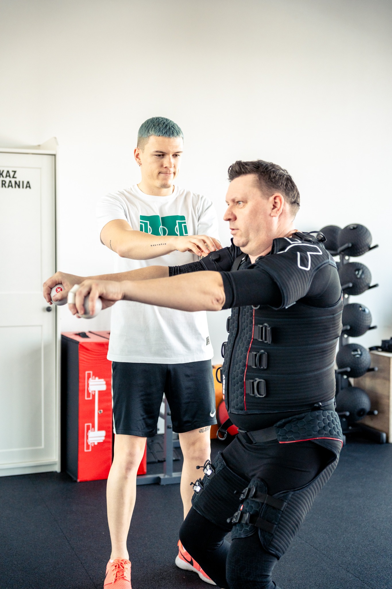 Mężczyzna w czarnym kombinezonie EMS ćwiczy przysiady z pomocą trenera, który trzyma małe ciężarki.