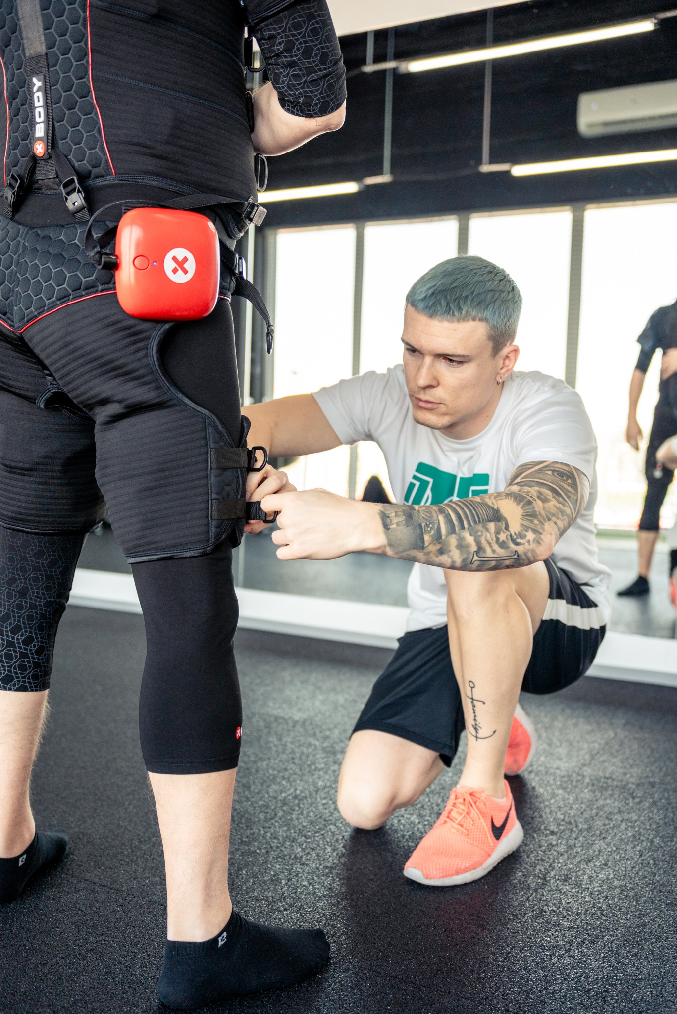 Mężczyzna w białej koszulce z tatuażami dopasowuje strój EMS do treningu elektrostymulacji na nodze klienta w czarnym kombinezonie z czerwonym urządzeniem.