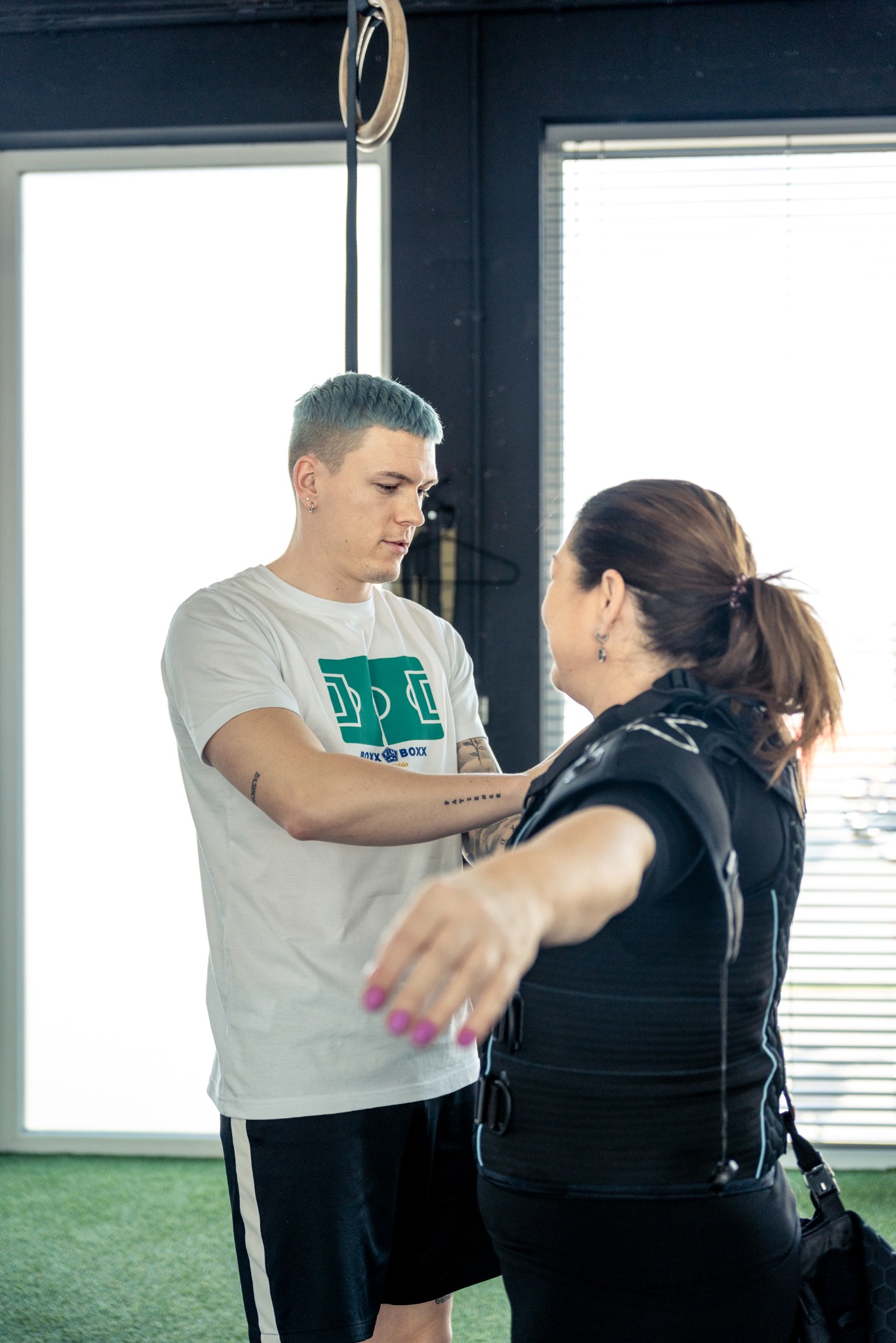 Mężczyzna z niebieskimi włosami w białej koszulce pomaga kobiecie założyć kamizelkę do elektrostymulacji mięśni w siłowni.