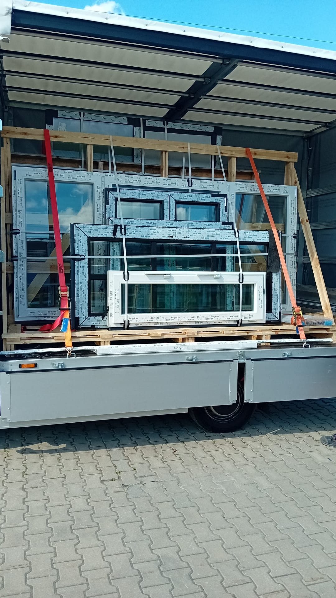 Okna PCV zabezpieczone pasami transportowymi na palecie wewnątrz naczepy busa, widoczne etykiety producenta SCHÜCO.