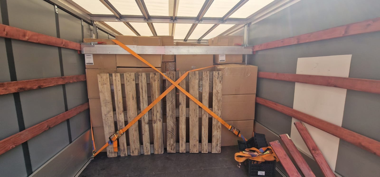Wnętrze busa załadowane kartonami, zabezpieczone paletami i pomarańczowymi pasami transportowymi, widoczne drewniane listwy na ścianach i metalowa konstrukcja dachu.
