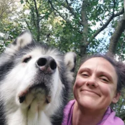 Selfie kobiety w fioletowej bluzie z kapturem i dużego, czarno-białego psa rasy malamut na tle drzew.