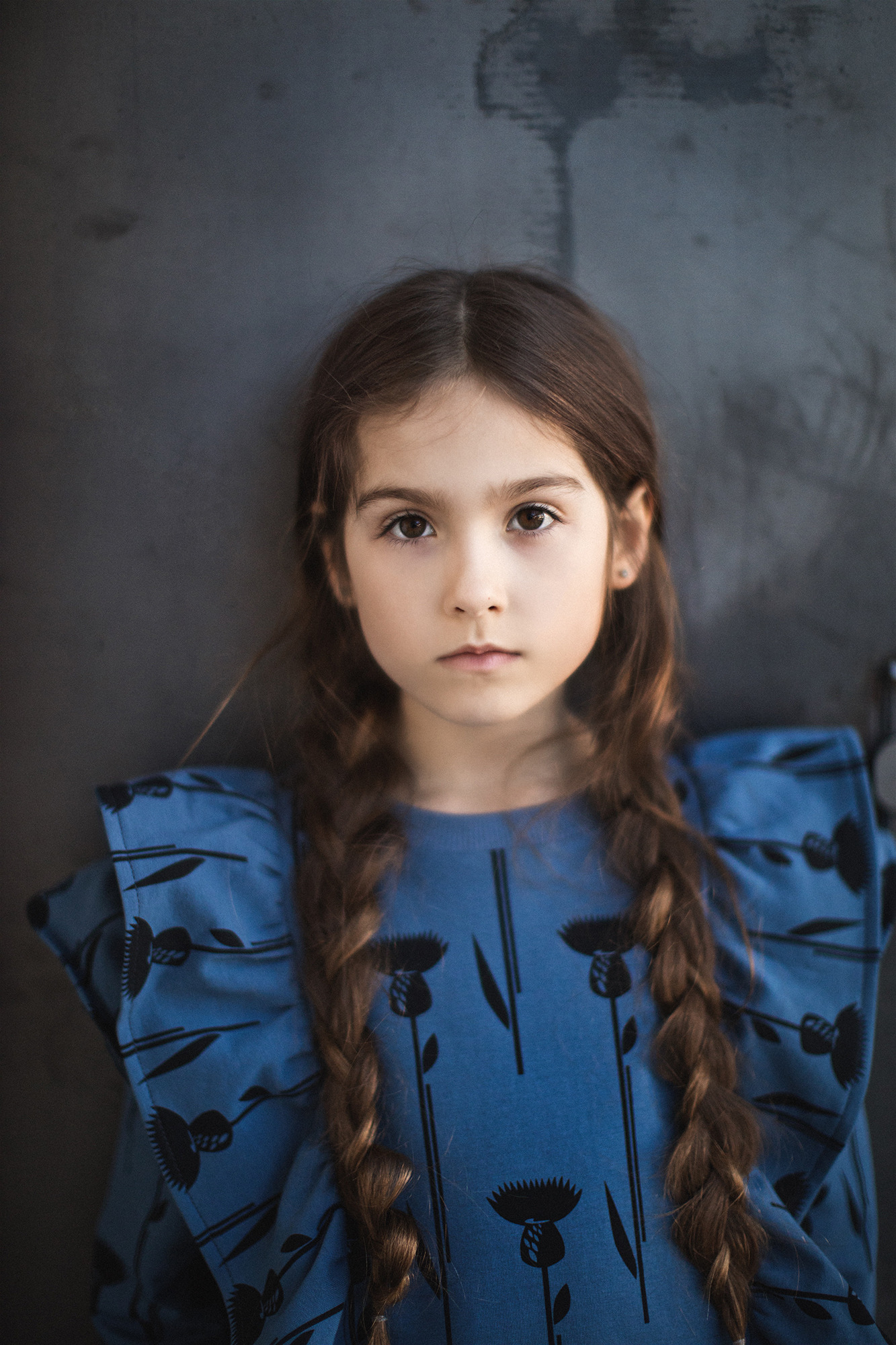 Portret dziewczynki z brązowymi warkoczami w niebieskiej sukience z motywem ostu, na tle szarej ściany.