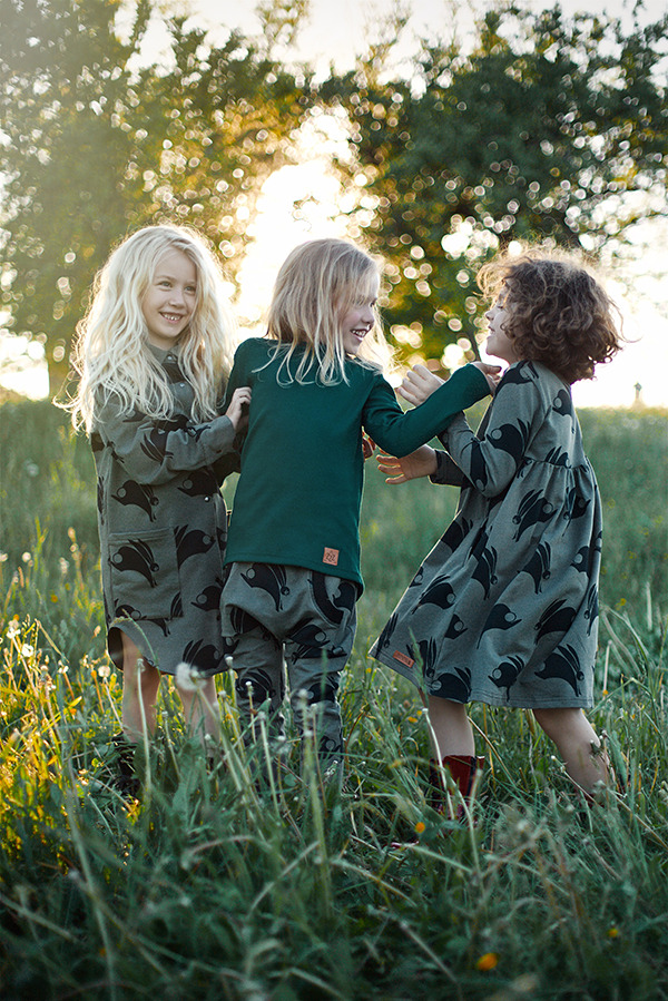 Trzy uśmiechnięte dziewczynki w autorskich ubraniach z motywem zająca, stojące na łące w promieniach zachodzącego słońca.