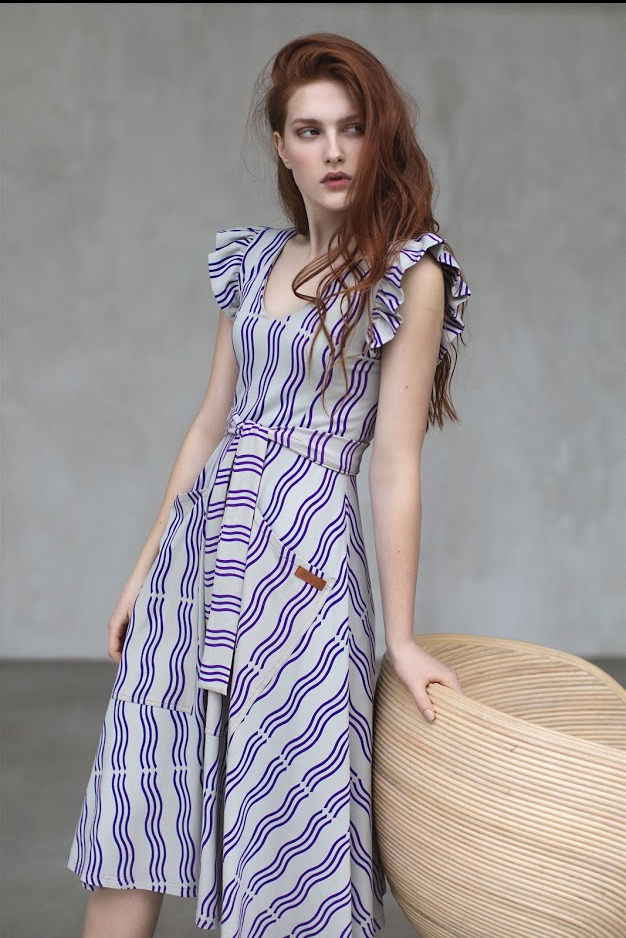 Modelka w letniej sukience w biało-fioletowe paski z falbankami i paskiem w talii, opierająca się o beżowy fotel.