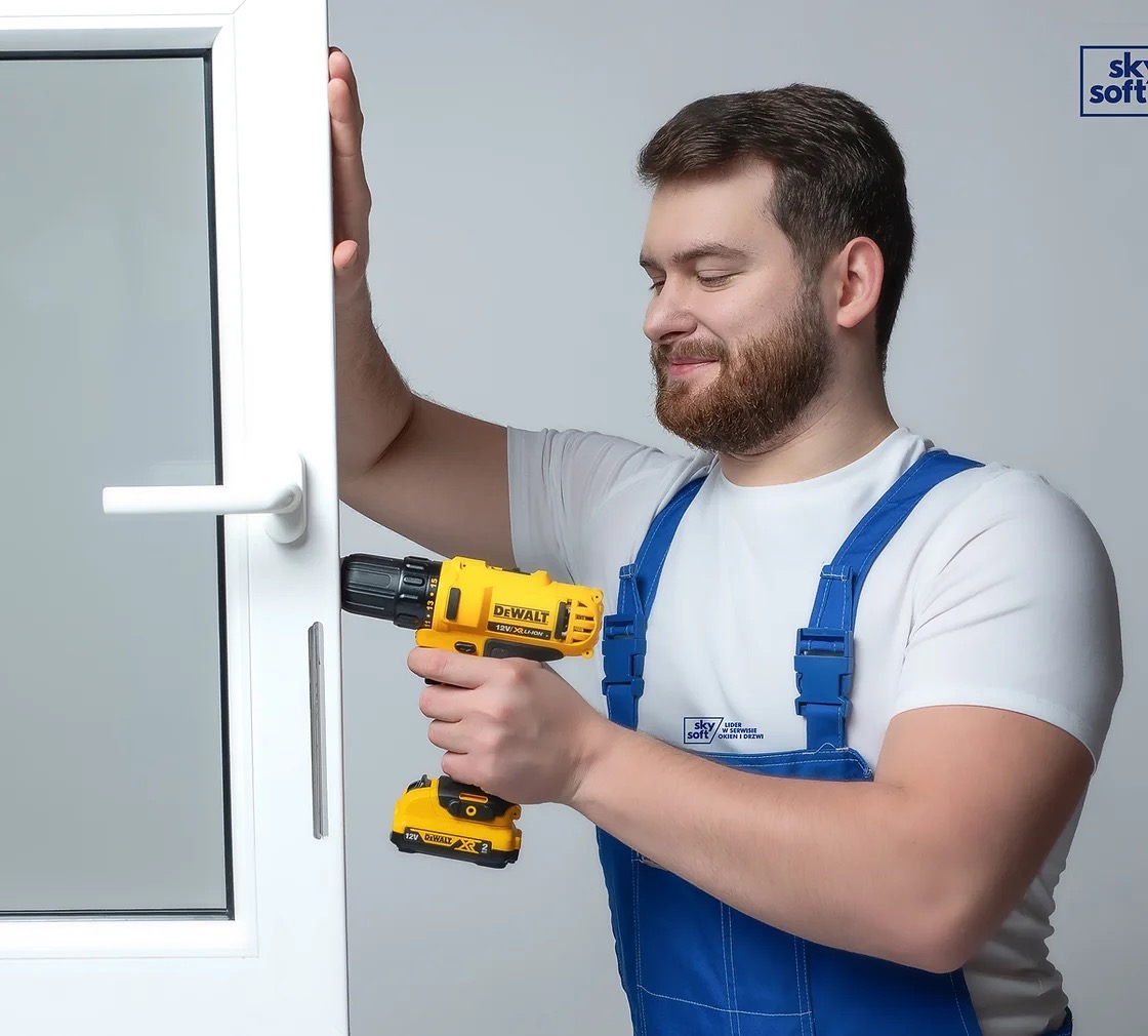 Mężczyzna w niebieskim kombinezonie, z uśmiechem, używa żółtej wkrętarki DeWalt do naprawy białego okna z widoczną klamką i logiem firmy Sky Soft na kombinezonie.