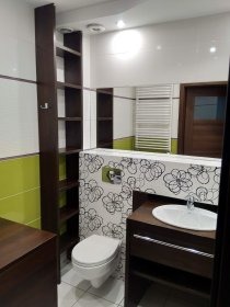 Nowoczesna łazienka z białymi płytkami przełamanymi poziomym pasem zieleni, toaleta z wzorem kwiatowym, ciemnobrązowe szafki pod umywalką i wysoki regał z półkami. Widoczne białe grzejniki...