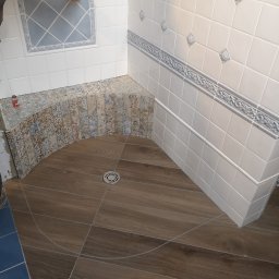 Usługi ogulnobudowlane - Fragment łazienki w trakcie prac wykończeniowych; widoczne ułożone płytki podłogowe imitujące drewno, fragment brodzika z mozaiką oraz białe płytki ścienne z dekoracyjnym pasem i ozdobnymi...
