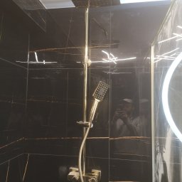 Usługi ogulnobudowlane - Kabina prysznicowa z czarnymi płytkami imitującymi marmur ze złotymi żyłami, złota armatura, deszczownica sufitowa i oświetlenie LED w lustrze odbijające fotografa.