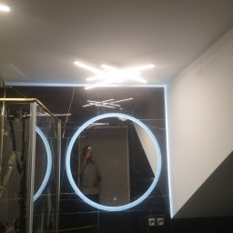 Usługi ogulnobudowlane - Nowoczesna łazienka z czarnymi płytkami imitującymi marmur, okrągłym lustrem z podświetleniem LED, złotą armaturą i kabiną prysznicową ze złotymi profilami, oświetlona lampą sufitową w kształcie...