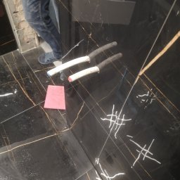 Usługi ogulnobudowlane - Czarna, błyszcząca płytka podłogowa z złotymi żyłkami, z widocznymi śladami po fugowaniu, różowa gąbka do czyszczenia i fragment instalacji hydraulicznej z rurami PCV