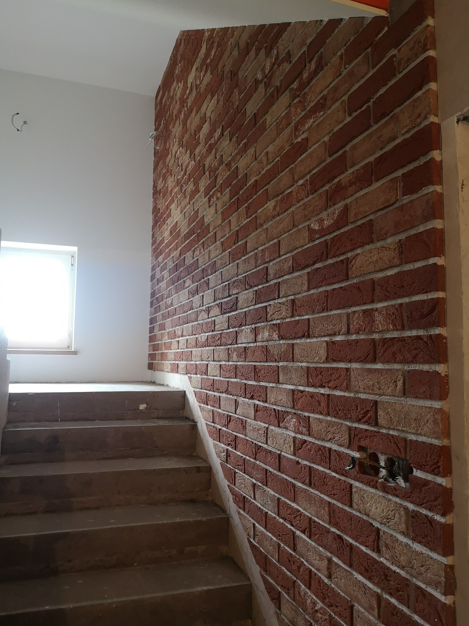 Wykończenie klatki schodowej z ceglaną ścianą w odcieniach czerwieni i beżu, widoczne schody w surowym stanie i wystające przewody elektryczne.