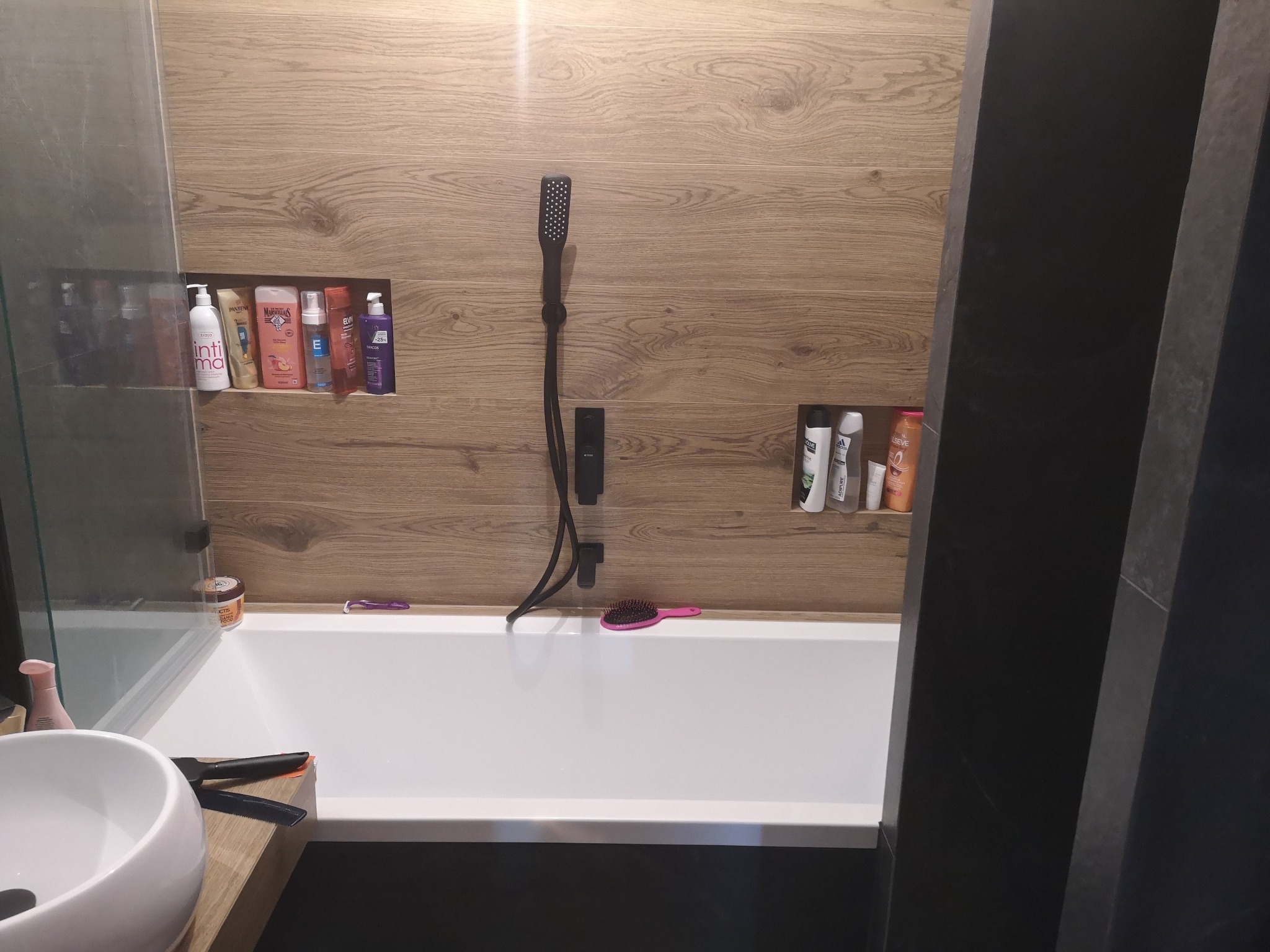 Nowoczesna łazienka z drewnopodobnymi płytkami, czarną armaturą i wnękami na kosmetyki, widok na wannę z akcesoriami.