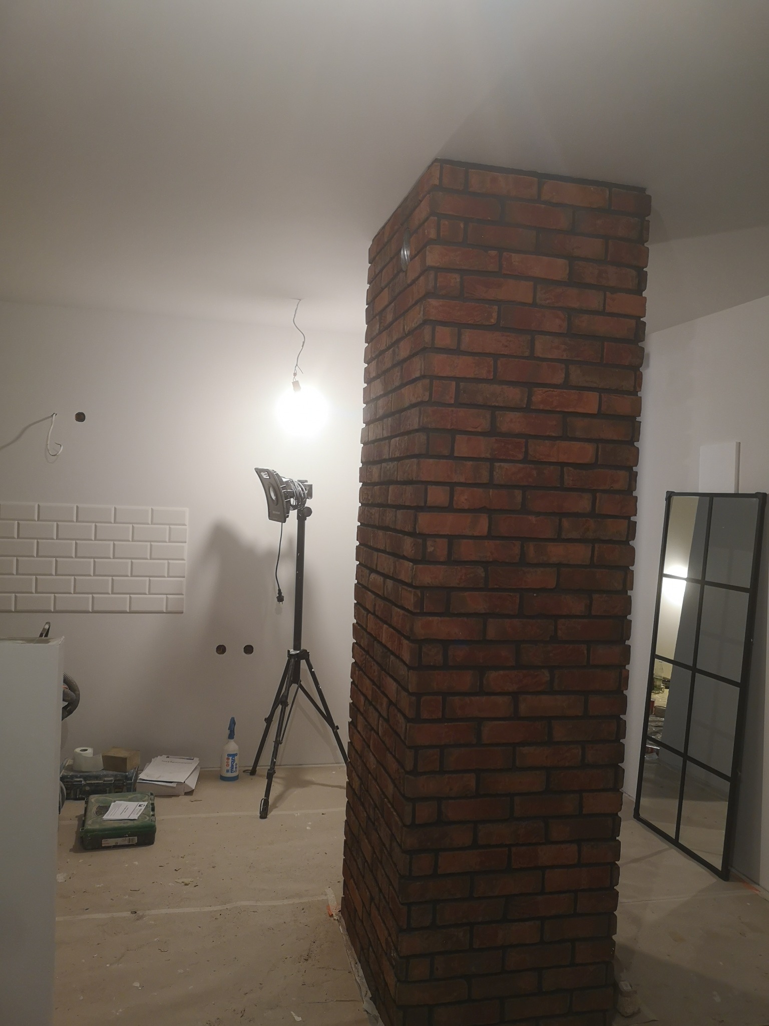 Kolumna wykończona czerwoną cegłą w surowym wnętrzu, widoczna lampa na statywie, fragment ułożonej glazury na ścianie i lustro w czarnej ramie.