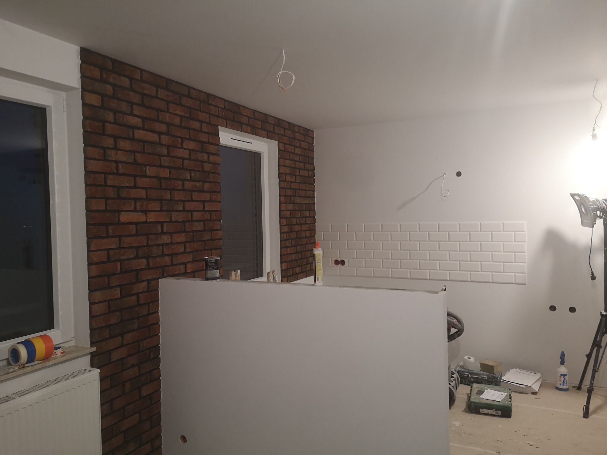 Wnętrze w trakcie prac wykończeniowych: ściana z cegły klinkierowej, fragment ściany z białymi płytkami typu metro, widoczne narzędzia i materiały budowlane, surowe kable wystające z sufitu.