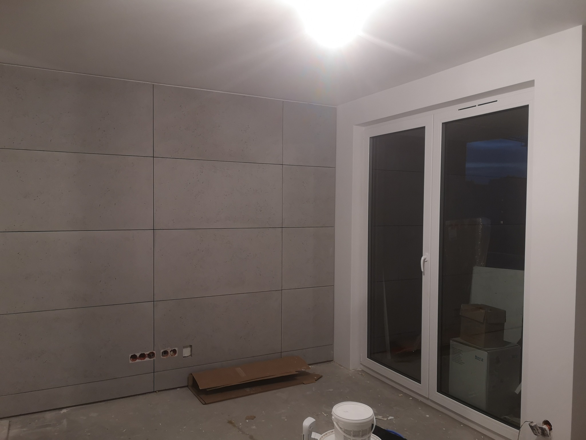 Ściana wyłożona szarymi płytami imitującymi beton, z widocznymi puszkami elektrycznymi, białe okno balkonowe, podłoga w trakcie prac wykończeniowych.