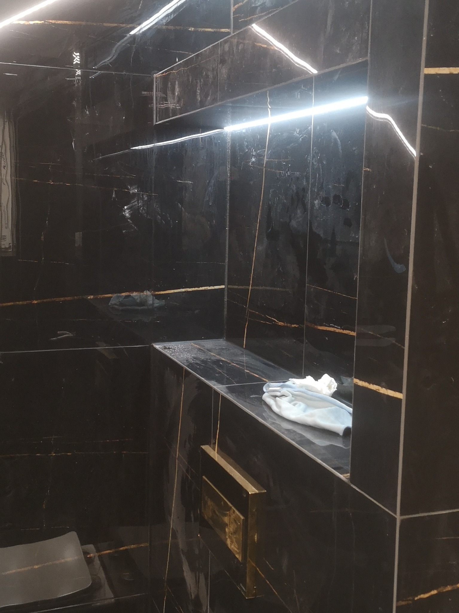 Czarna łazienka z marmurowymi płytkami z żyłami złota, podświetlona półka i fragment toalety w dolnym lewym rogu, na półce leży biały ręcznik.