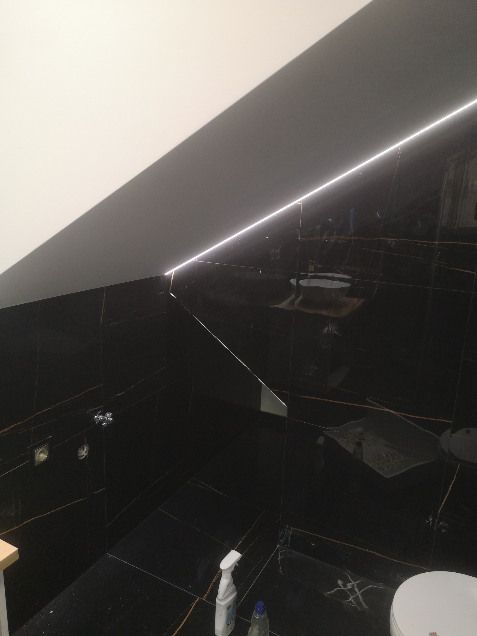 Ukończona łazienka z czarnymi, marmurkowymi płytkami, oświetleniem LED wzdłuż skośnego sufitu, widoczne gniazdka elektryczne, fragment toalety i butelka środka czyszczącego na podłodze.